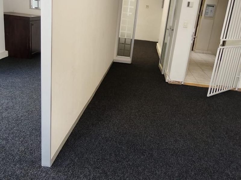An empty office space featuring dark carpeting, partition walls, exposed ducting, and a security gate, offering a functional layout for professional use.