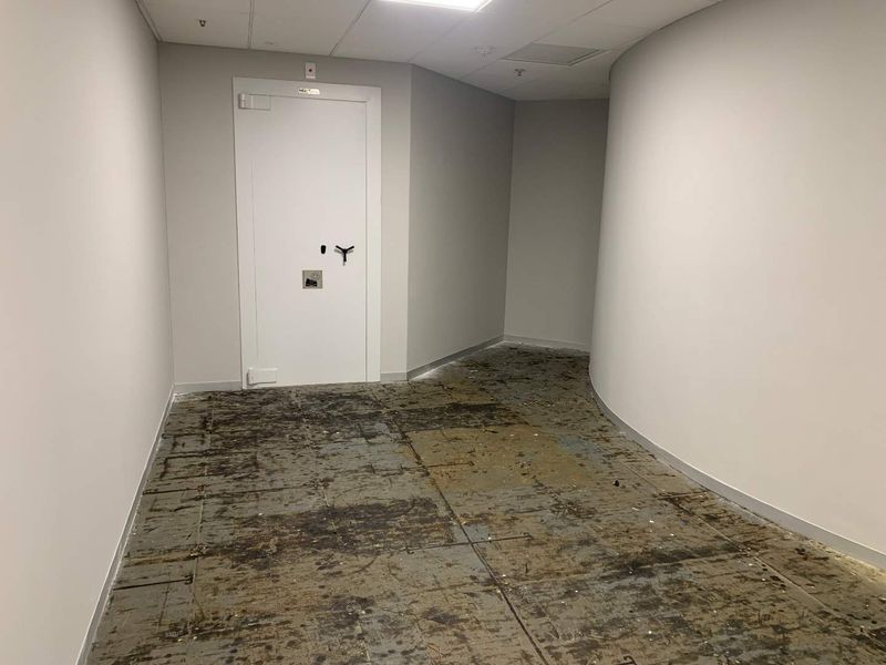 A vacant office hallway featuring a curved wall, suspended ceiling with recessed lighting, and a fire-rated door, currently requiring new flooring installation.