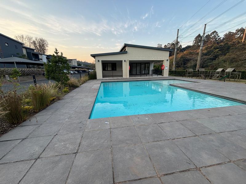A well-maintained communal swimming pool area featuring a paved deck, landscaped surroundings, and an adjacent clubhouse building within the apartment complex.