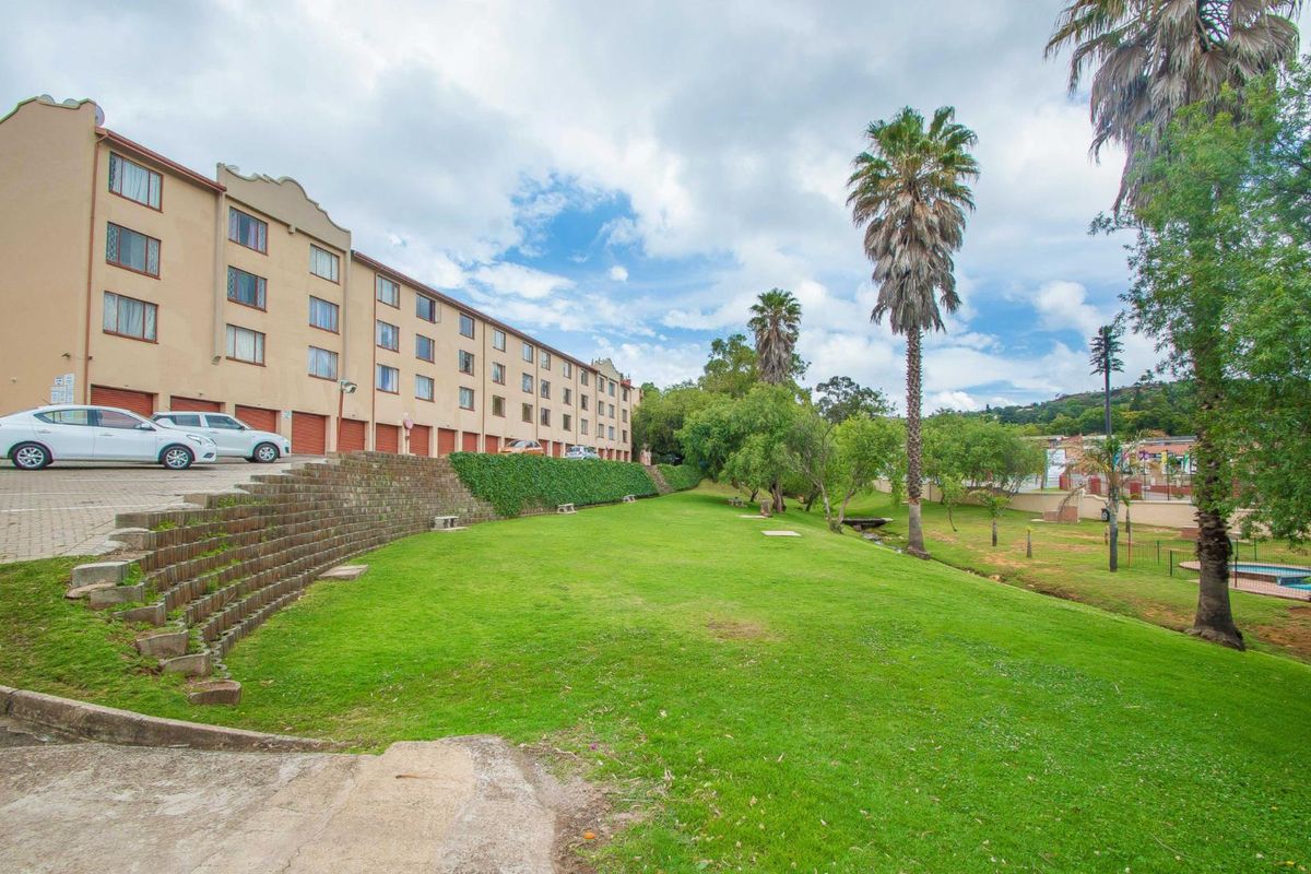 Exterior view of the apartment complex featuring a large communal lawn, mature trees, and a paved parking area with a retaining wall.