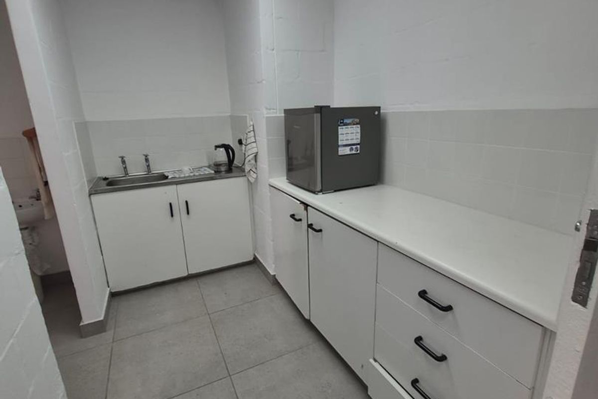 A functional office kitchenette featuring built-in white cabinetry, a stainless steel sink, tiled splashbacks, and durable floor tiles.