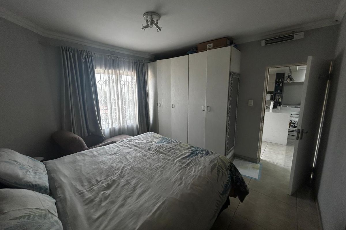A bedroom featuring built-in wardrobes, tiled flooring, and a window with security bars, offering a view into the adjacent kitchen area.