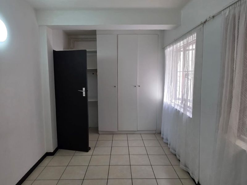 A clean, empty bedroom featuring tiled flooring, built-in cupboards, and large windows with curtains, offering a neutral space ready for personalization.