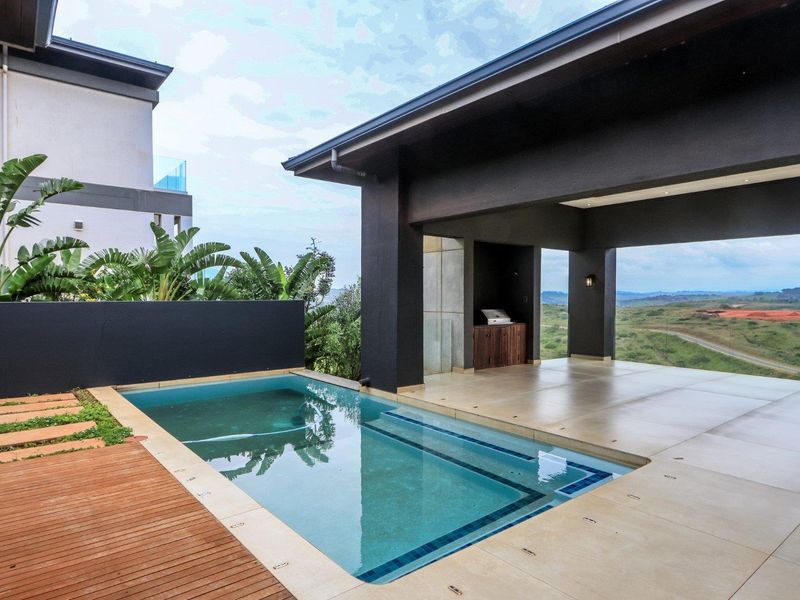 Modern covered patio area featuring a swimming pool, timber decking, and a built-in braai, offering expansive views of the surrounding landscape.