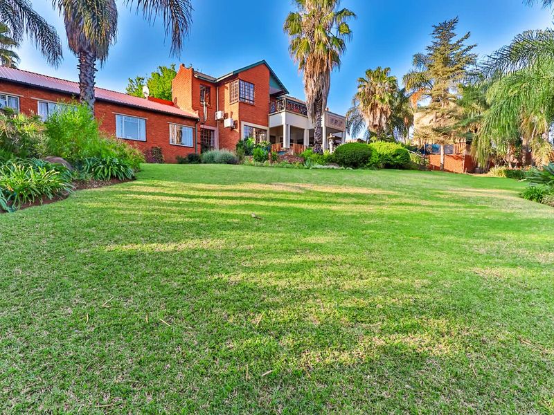 A view of the rear elevation of the brick house, featuring a spacious lawn, mature palm trees, and an upper-level balcony.