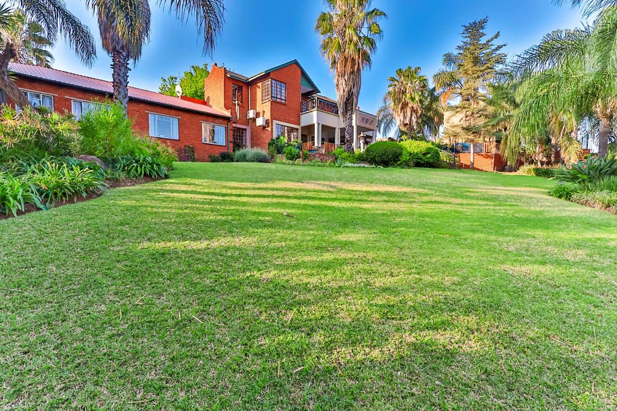 A view of the rear elevation of the brick house, featuring a spacious lawn, mature palm trees, and an upper-level balcony.