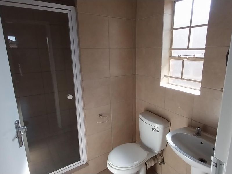 A functional bathroom featuring a shower enclosure, pedestal basin, and toilet, finished with neutral wall tiling and a window for natural light.