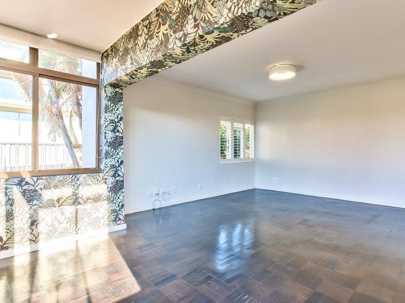 A bright and spacious living area featuring dark parquet flooring, large windows with plantation shutters, and a decorative patterned feature wall.