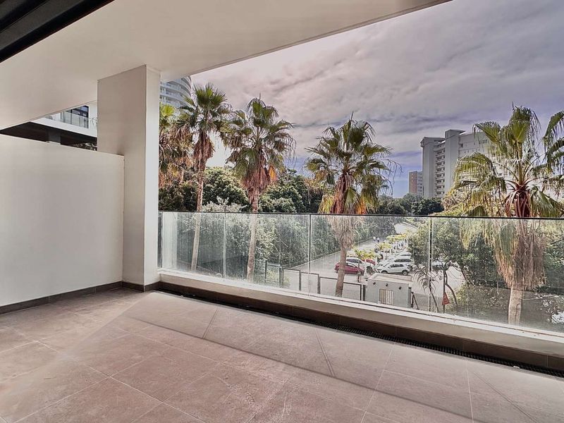 A modern covered balcony featuring tiled flooring and a frameless glass balustrade, offering an elevated view of the surrounding area and palm trees.