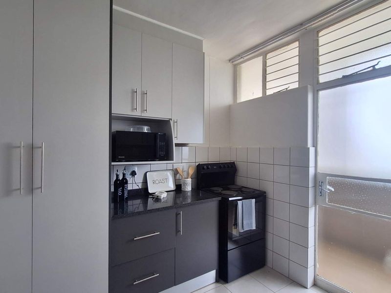 A functional kitchen featuring modern grey cabinetry, granite countertops, a tiled backsplash, and a freestanding stove, with natural light from louvre windows.