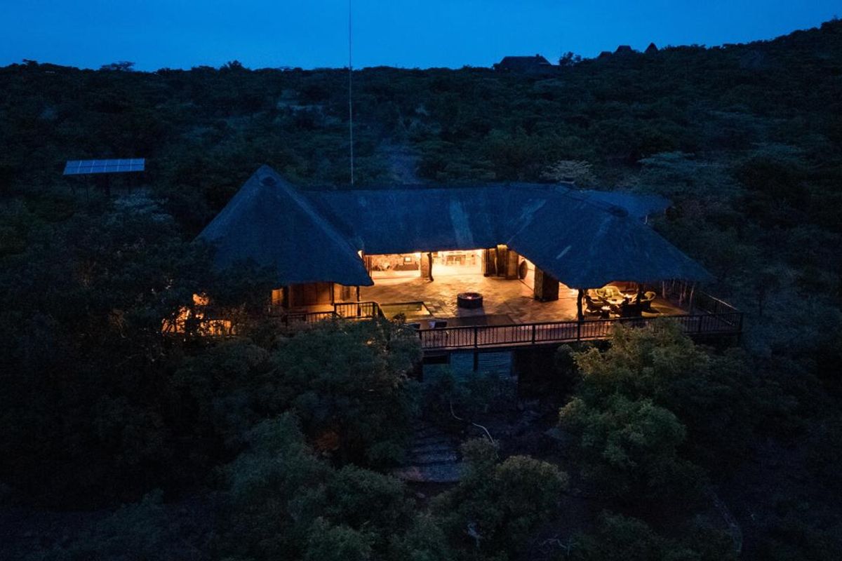 An elevated aerial view of a thatched-roof farm lodge at dusk, featuring an expansive wooden deck and surrounding natural bushveld landscape.