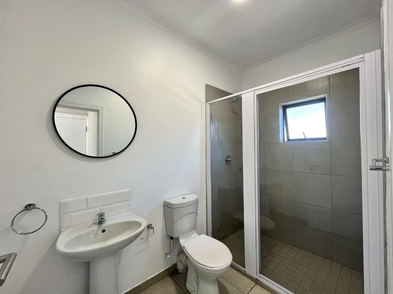 A clean, modern bathroom featuring a pedestal washbasin, toilet, and a glass-enclosed shower with neutral wall tiling.