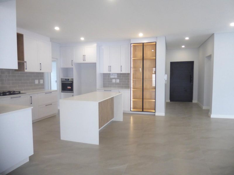 Modern, open-plan kitchen featuring white cabinetry, a central island, built-in oven, gas hob, and elegant glass-front display shelving with integrated lighting.