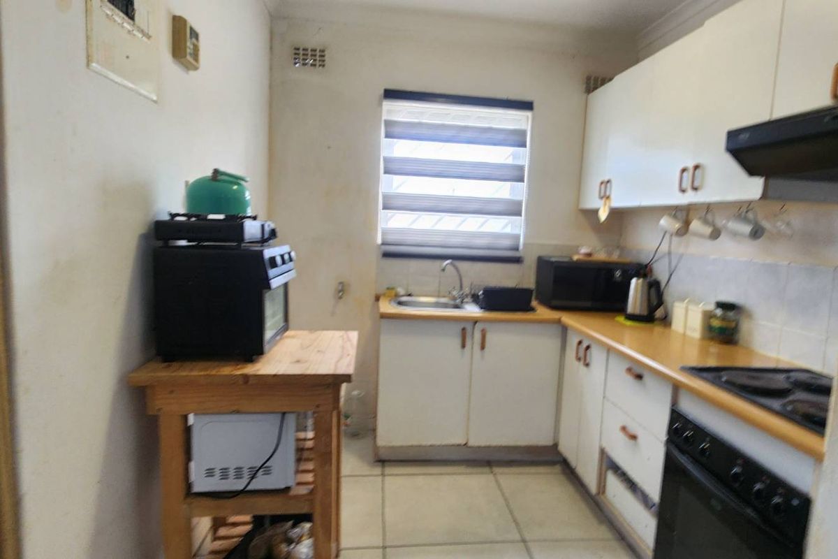 A compact kitchen featuring white cabinetry, laminate countertops, integrated oven and hob, and tiled flooring, currently showing signs of wear and dated finishes.