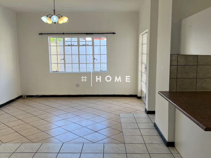 An open-plan living and kitchen area featuring neutral tiled flooring, a breakfast counter, and natural light through a window with security bars.