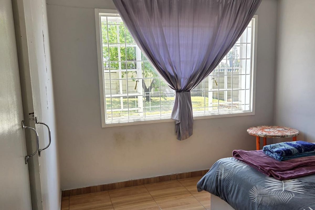 A bedroom featuring tiled flooring and a window fitted with security bars, offering a view of the exterior garden area.