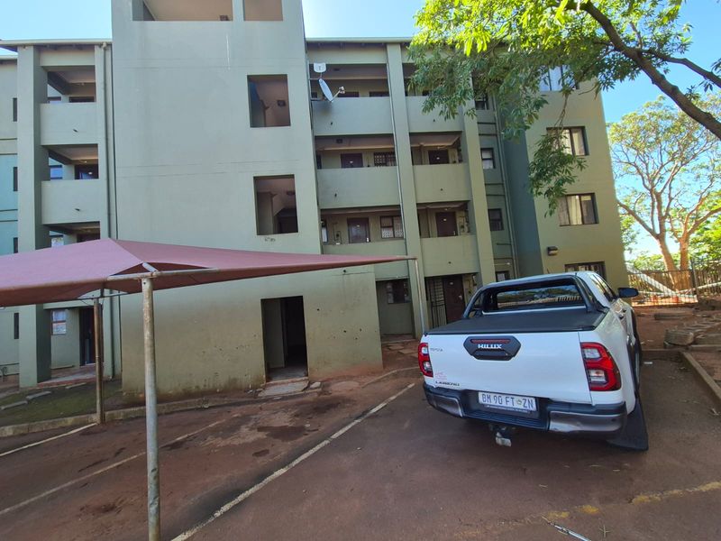 Exterior view of a multi-story apartment building featuring a paved driveway and a shade cloth carport structure.
