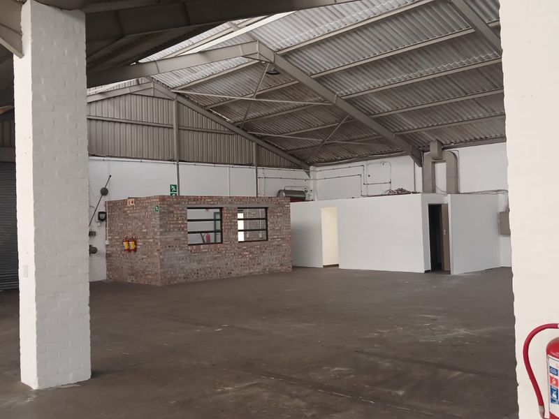 Spacious industrial warehouse unit featuring high ceilings with exposed steel trusses, concrete flooring, a central brick office partition, and a large roller shutter door.