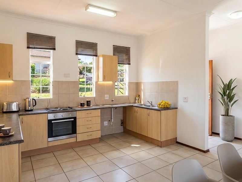 A functional kitchen featuring light wood cabinetry, dark laminate countertops, built-in oven, electric hob, and neutral tiled flooring with ample natural light.