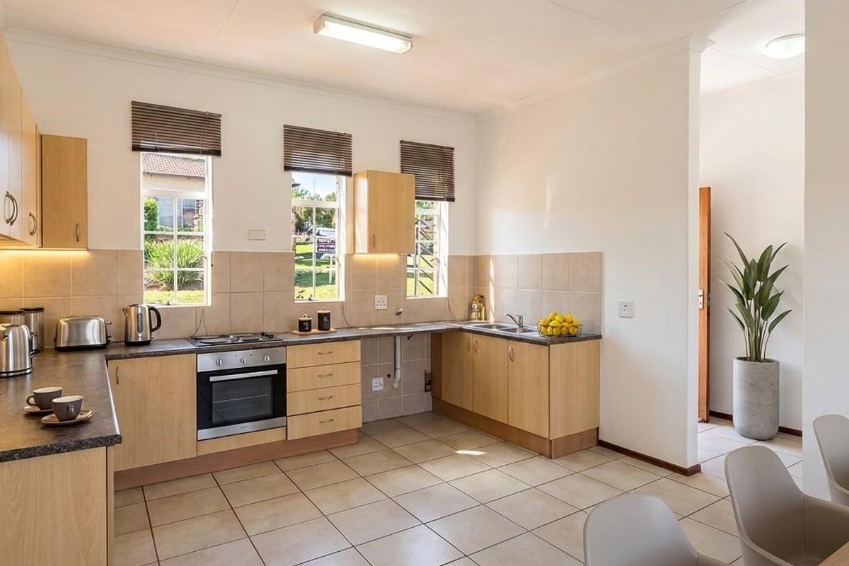A functional kitchen featuring light wood cabinetry, dark laminate countertops, built-in oven, electric hob, and neutral tiled flooring with ample natural light.