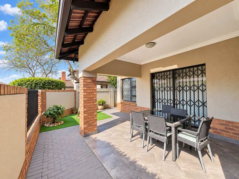 A covered patio area featuring tiled flooring, brick support pillars, and a secure sliding door entrance, overlooking a small private garden space.