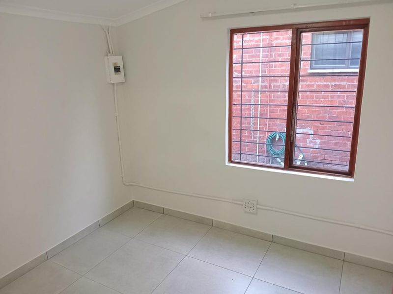 A clean, empty bedroom featuring neutral tiled flooring, a timber-framed window with security bars, and visible surface-mounted electrical infrastructure.
