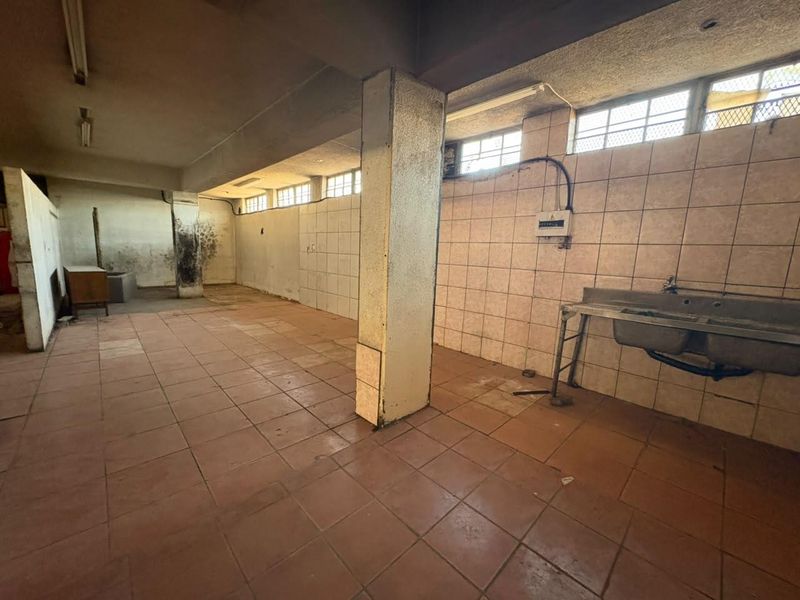 A spacious, vacant utility area featuring tiled walls and flooring, a stainless steel double sink, and exposed structural columns, requiring significant refurbishment.