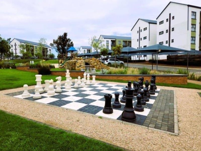 A well-maintained communal outdoor area featuring a large garden chess set, landscaped grounds, and shaded parking structures within the residential estate.