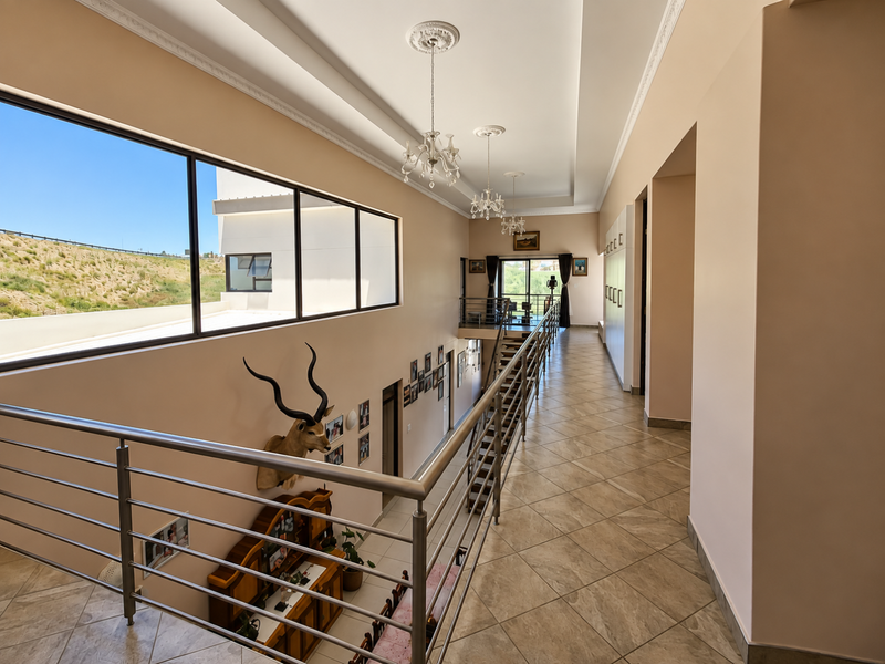 Spacious upper-level hallway featuring tiled flooring, stainless steel balustrades, and large windows overlooking the exterior, complemented by elegant chandeliers and recessed ceiling details.