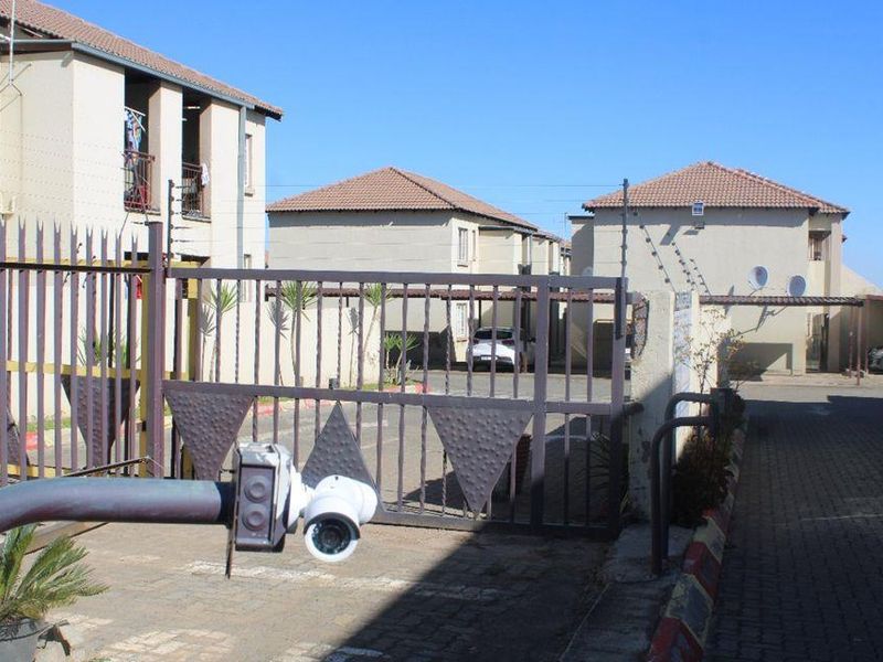 Front view of the townhouse complex entrance featuring a security gate, electric fencing, and paved driveway access to the parking area.