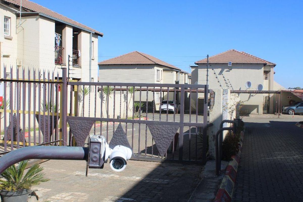 Front view of the townhouse complex entrance featuring a security gate, electric fencing, and paved driveway access to the parking area.