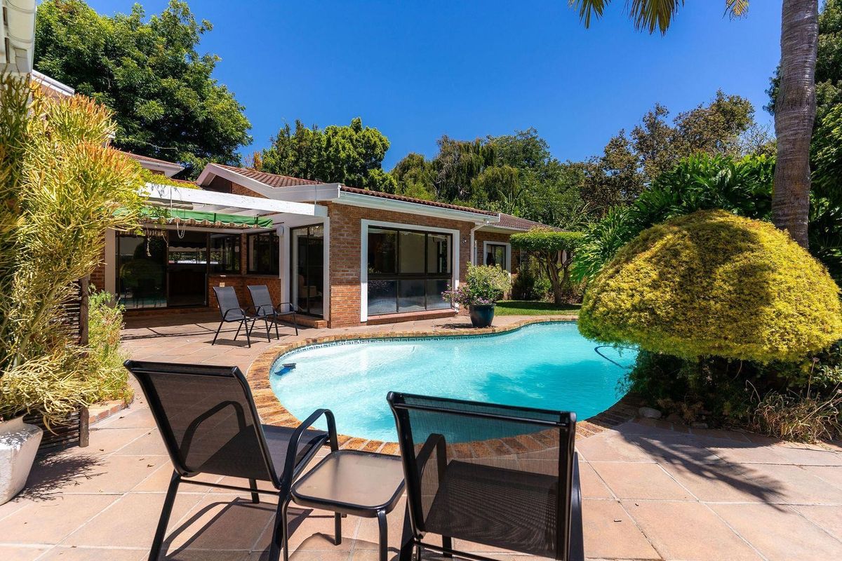 A well-maintained rear exterior featuring a paved patio area, a swimming pool, and established garden landscaping adjacent to the brick residence.