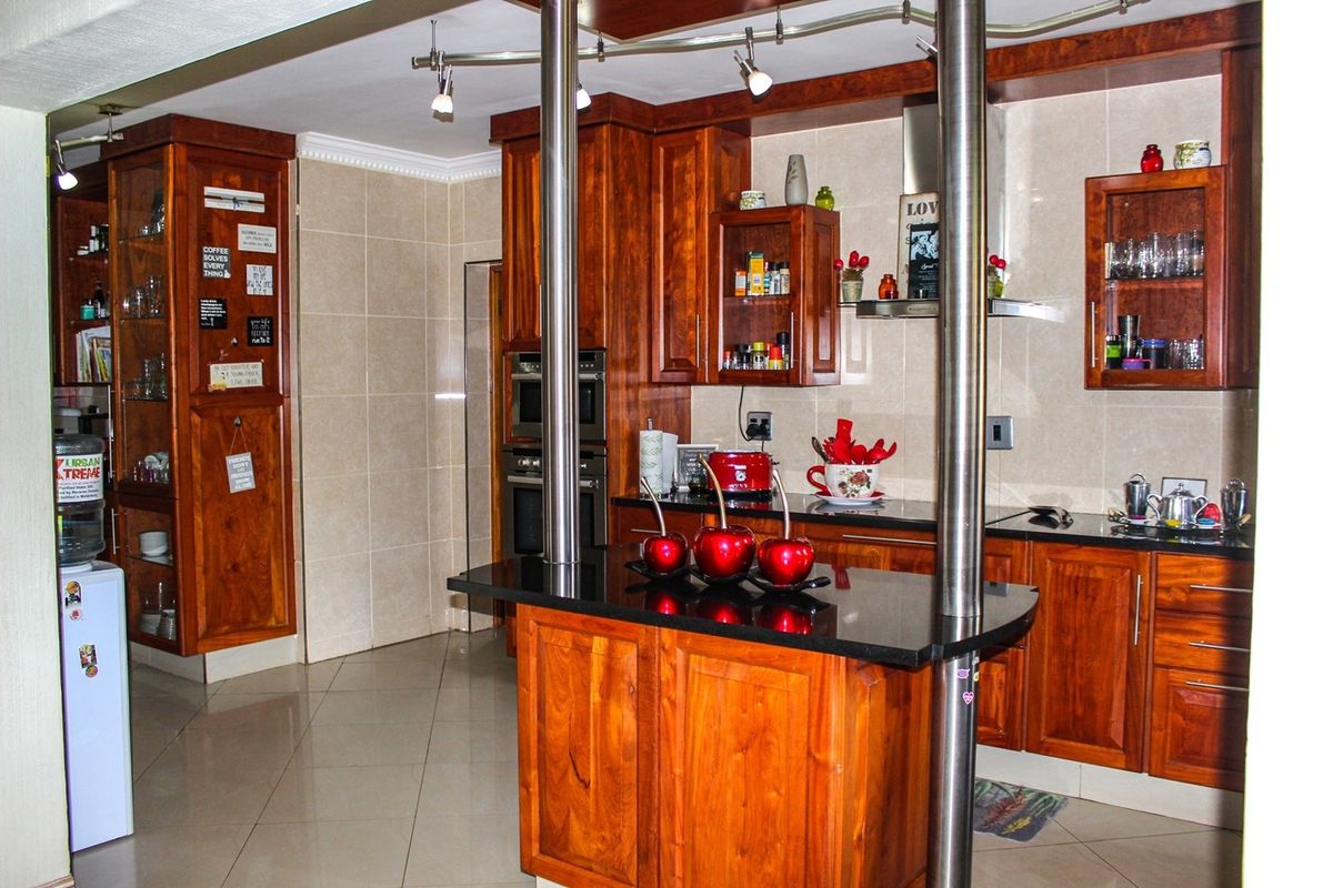 A kitchen featuring warm-toned wooden cabinetry, black granite countertops, a central island with stainless steel support pillars, and tiled flooring.