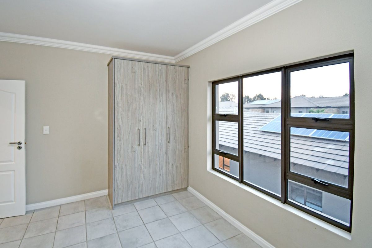 A bright, empty bedroom featuring neutral tiled flooring, built-in cupboards with a wood-grain finish, and large aluminium-framed windows.
