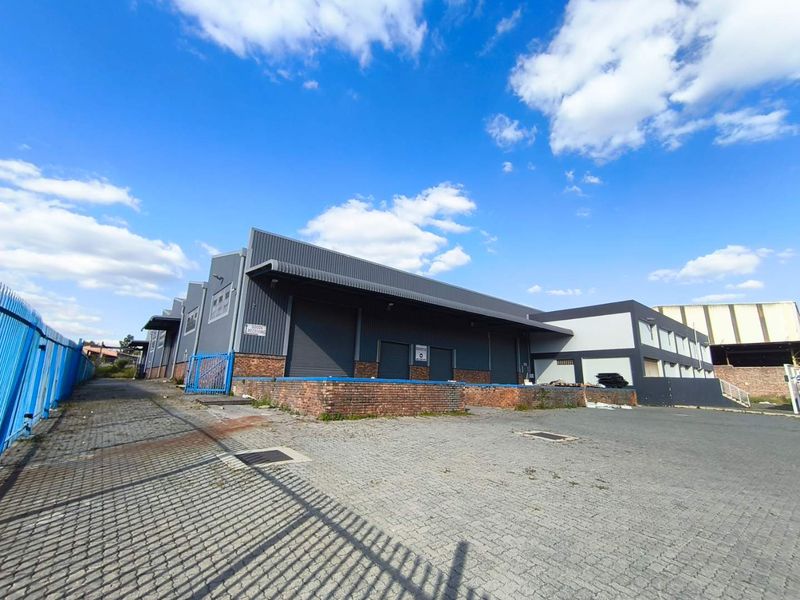 Industrial warehouse facility featuring multiple roller shutter doors, a raised loading dock, and a paved yard area enclosed by security fencing.