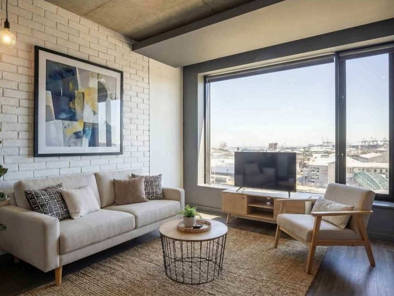 Modern living room featuring an exposed concrete ceiling, painted brick feature wall, and large windows offering expansive urban views.
