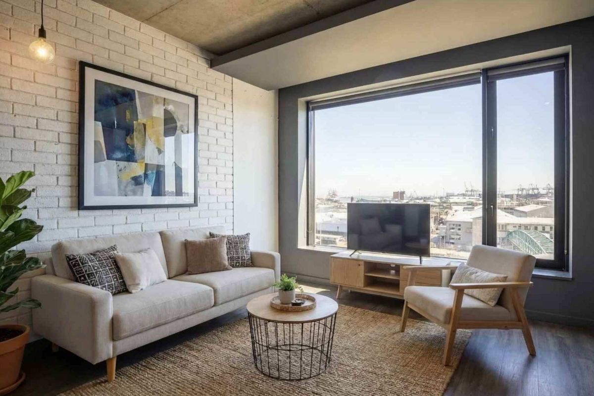 Modern living room featuring an exposed concrete ceiling, painted brick feature wall, and large windows offering expansive urban views.