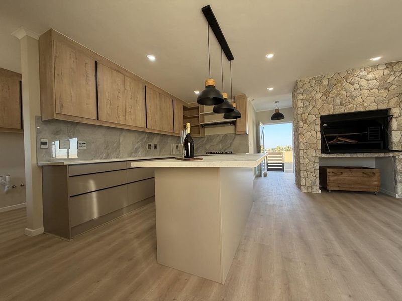 Modern open-plan kitchen featuring sleek cabinetry, a central island, and a striking stone-clad fireplace with a built-in braai, leading to an outdoor area.