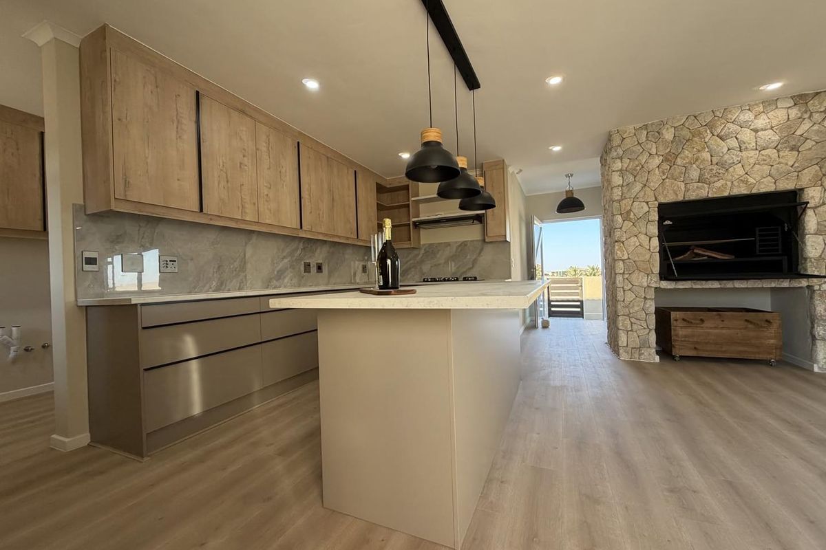 Modern open-plan kitchen featuring sleek cabinetry, a central island, and a striking stone-clad fireplace with a built-in braai, leading to an outdoor area.