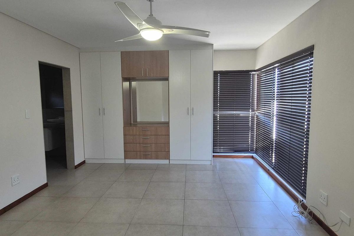 Spacious bedroom featuring built-in cupboards with integrated vanity, tiled flooring, ceiling fan, and large windows fitted with dark venetian blinds.