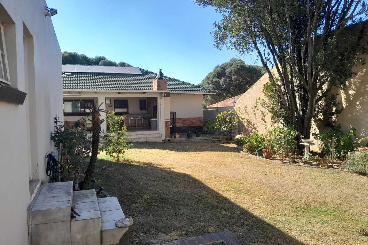Rear view of the property featuring a spacious lawn, a covered patio with a built-in braai, and a tiled roof with solar panels.