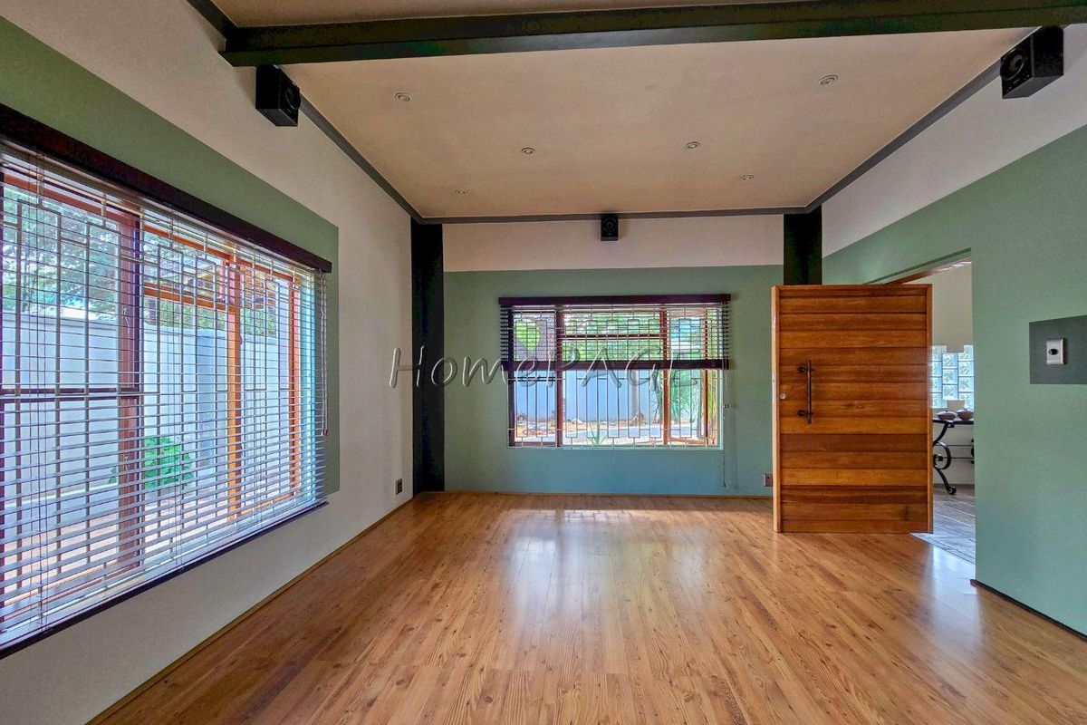 A spacious, light-filled living room featuring warm wood-look flooring, recessed ceiling lighting, and large windows with security bars.