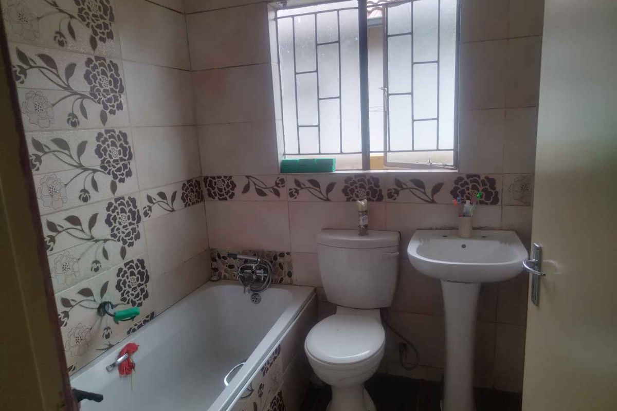 A compact bathroom featuring a standard bathtub, pedestal basin, and toilet, finished with dated floral-patterned wall tiles and a security-barred window.