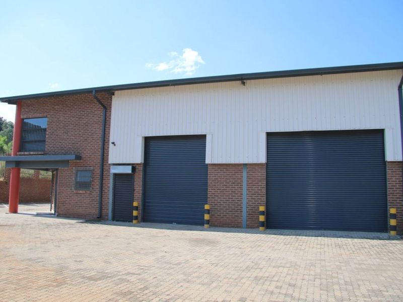 Industrial warehouse exterior featuring dual roller shutter doors, brick and metal cladding construction, and a paved driveway with protective security bollards.