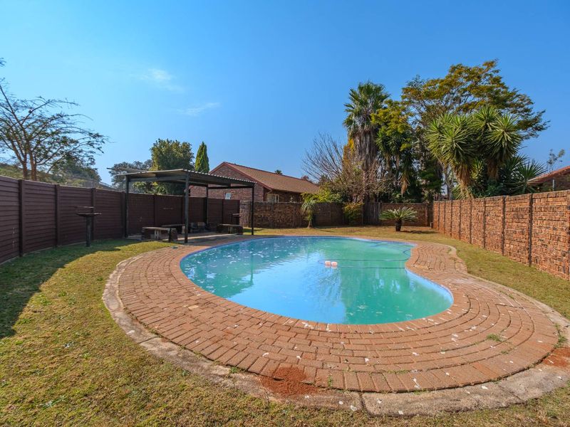 Outdoor pool area featuring a kidney-shaped swimming pool, brick paving surround, and a covered patio structure within a fenced garden setting.