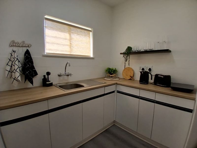 A modern, compact kitchen featuring white cabinetry with black accents, wood-look laminate countertops, a stainless steel sink, and a floating shelf.