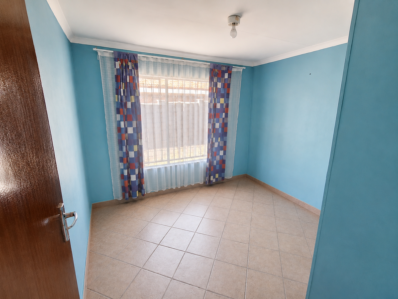 A bright, empty bedroom featuring neutral tiled flooring, blue painted walls, and a window fitted with security bars.