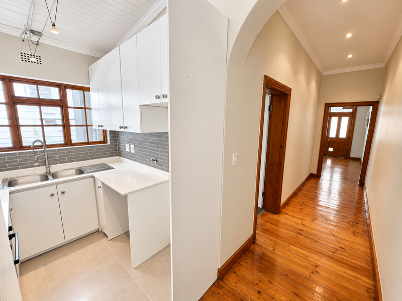 A bright, modern kitchen featuring white cabinetry, subway tile backsplash, and stainless steel sink, flowing into a hallway with polished hardwood floors.