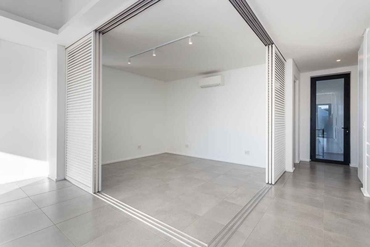 A bright, modern, and empty living space featuring large sliding louvered doors, neutral tiled flooring, and a wall-mounted air conditioning unit.