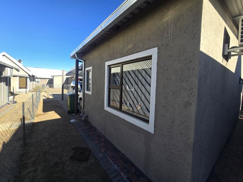 A view of the rear exterior of the apartment, featuring a textured plaster finish, aluminium windows with security bars, and a paved walkway.